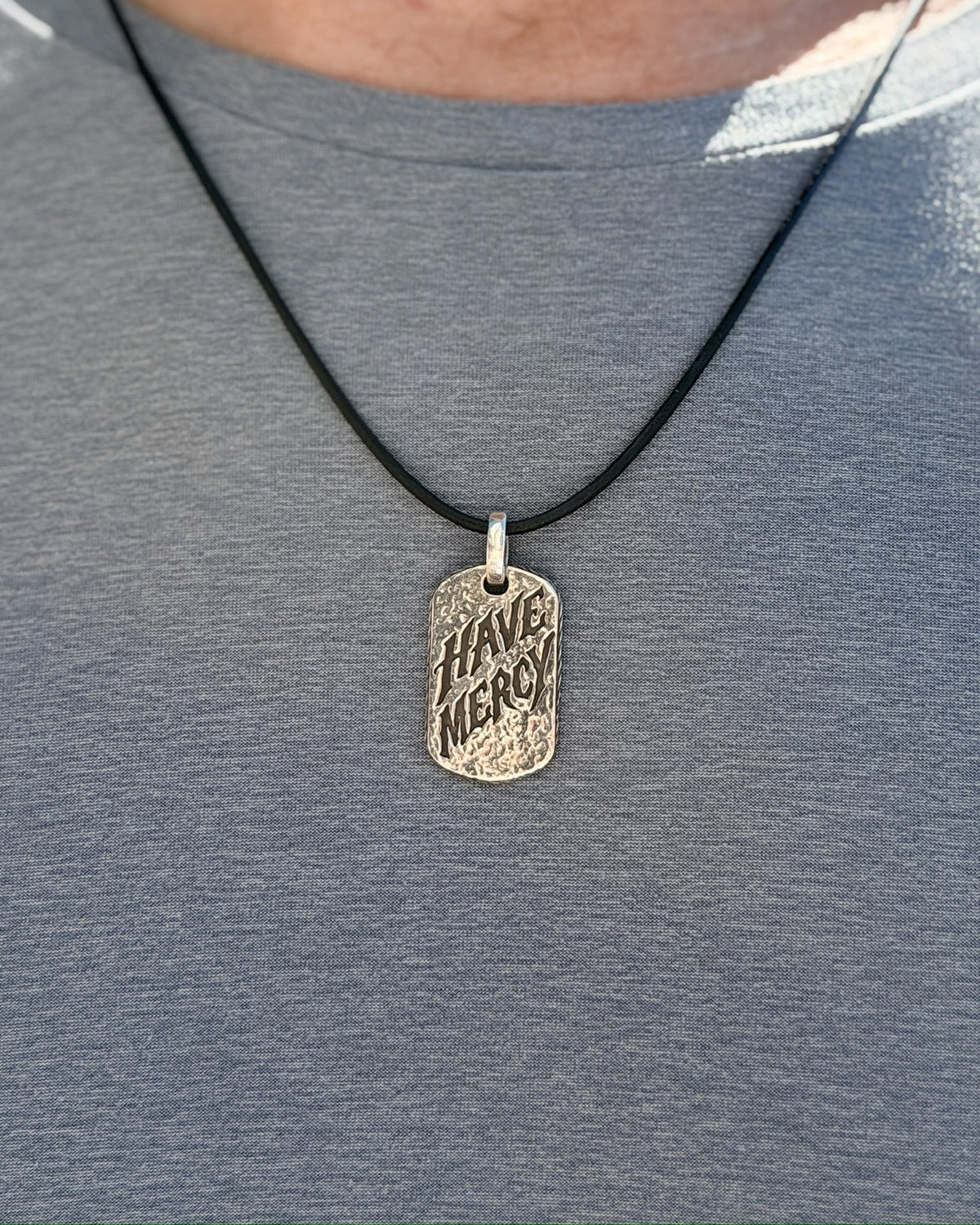 Man wearing the Have Mercy sterling silver pendant on a black leather cord over a gray t-shirt, front view